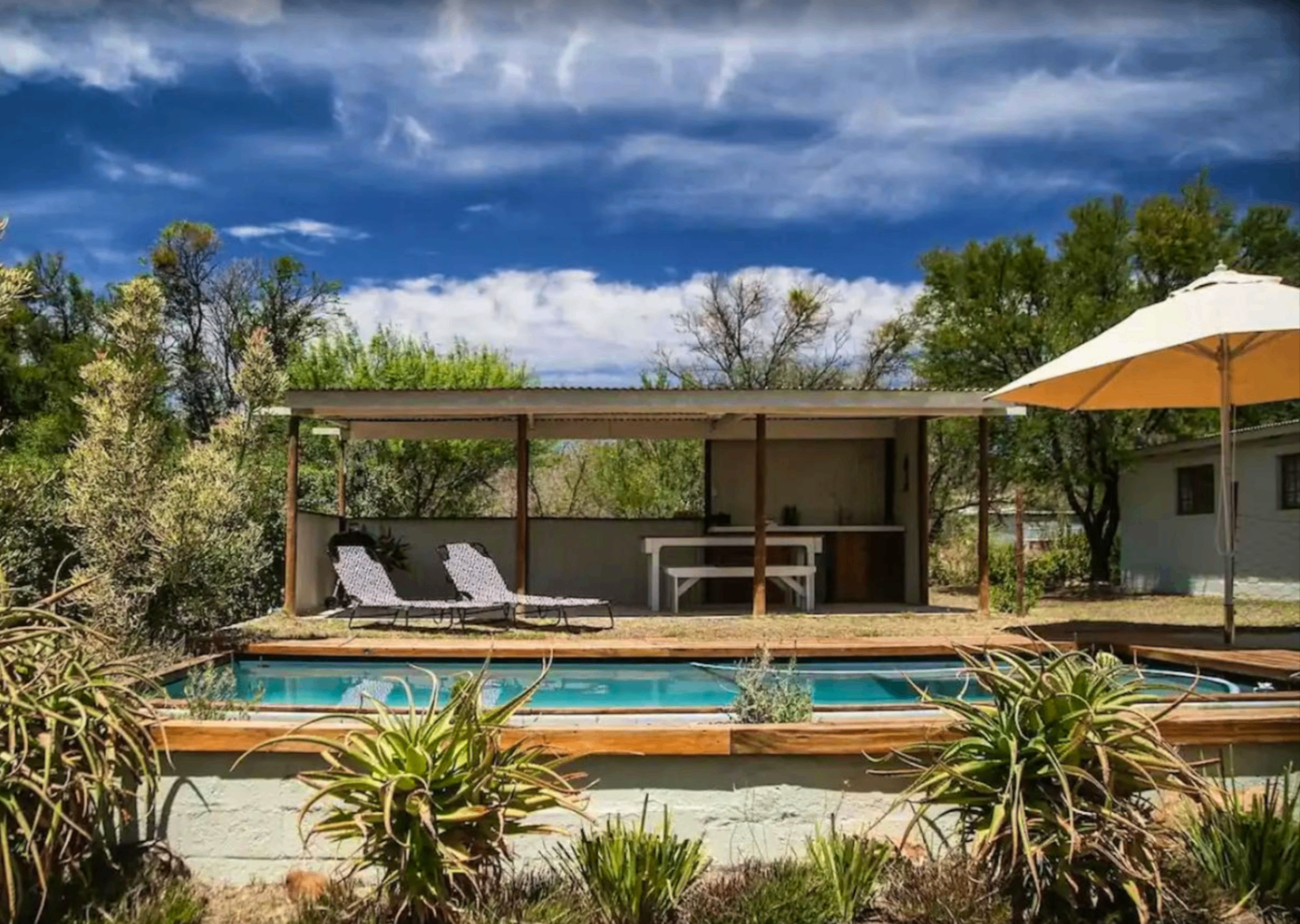Swimming pool and covered lapa at Karoo Feels with loungers, aloes, and dramatic Karoo sky.