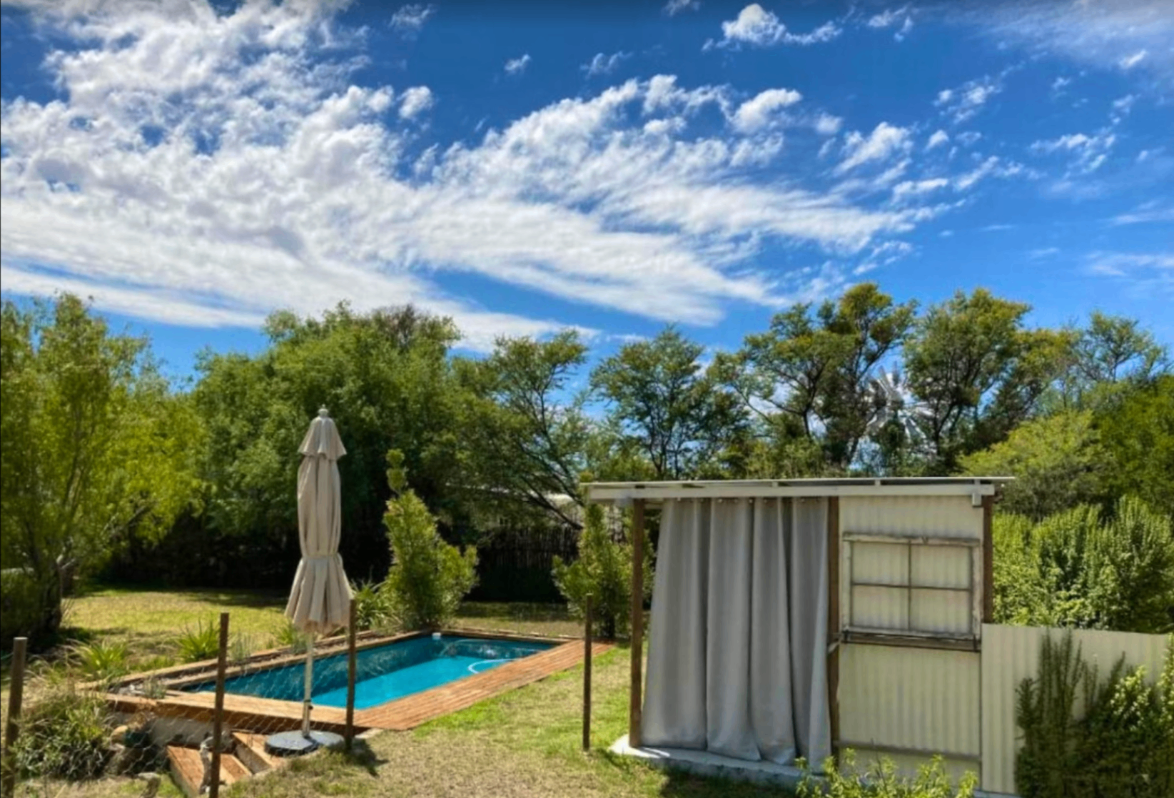 Garden pool at Karoo Feels on a bright day with umbrella, outdoor shower, and Karoo windmill in the distance.